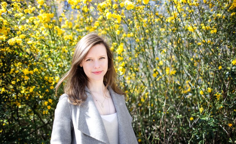 Eine junge Frau mit langen Haaren und einem lächelnden Gesichtsausdruck steht vor einem Hintergrund von leuchtend gelben Blumen.  | © privat