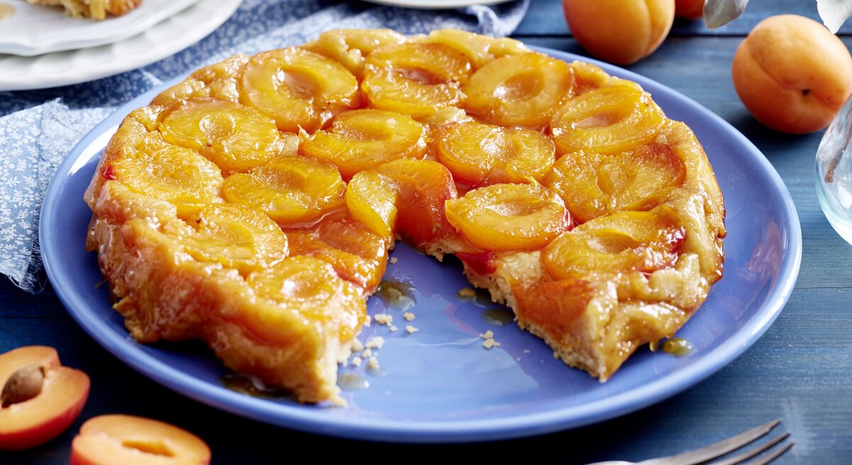 Aprikosen-Tarte-Tatin angeschnitten auf einer blauen Platte auf Holztisch mit Kaffeservice und ganzen Aprikosen