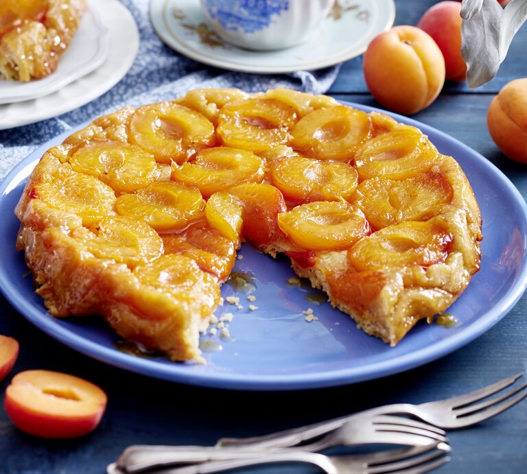 Aprikosen-Tarte-Tatin angeschnitten auf einer blauen Platte auf Holztisch mit Kaffeservice und ganzen Aprikosen | © Eat Club / Isabell Triemer