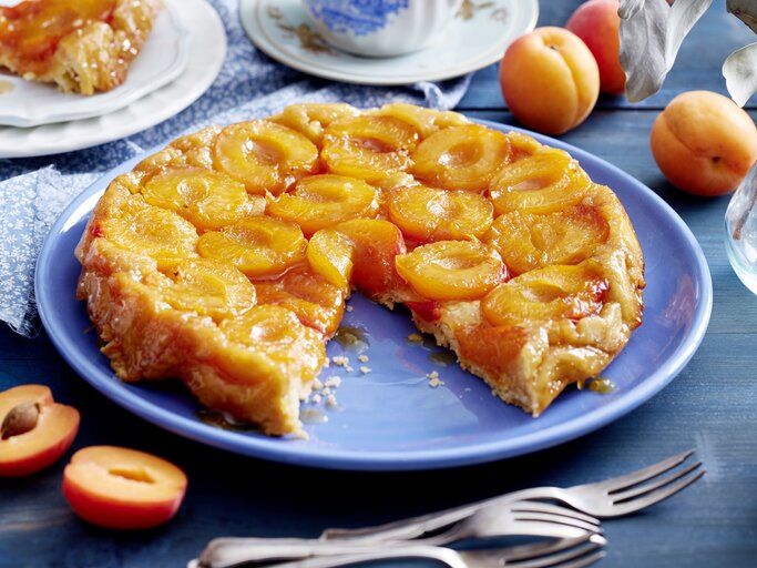 Aprikosen-Tarte-Tatin angeschnitten auf einer blauen Platte auf Holztisch mit Kaffeservice und ganzen Aprikosen | © Eat Club / Isabell Triemer