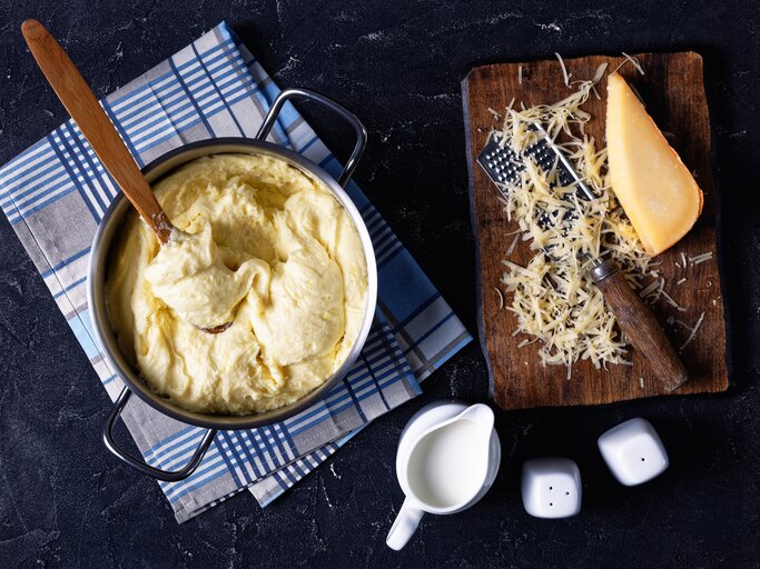 Käse-Kartoffelpüree in Topf auf dunkelblauem Untergrund. Daneben geriebener Käse. | © Shutterstock/from my point of view