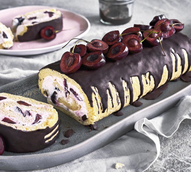 Minibiskuitrolle mit Kirschen und Schokoguss auf einer grauen Servierplatte auf einem grauen Tischtuch neben rosa Tellern auf einem grauen Steintisch.  | © StockFood / Springlane
