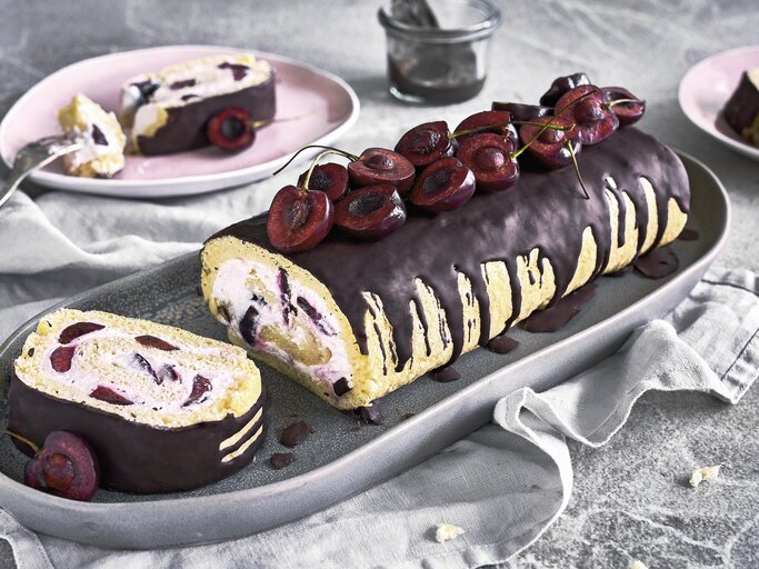 Minibiskuitrolle mit Kirschen und Schokoguss auf einer grauen Servierplatte auf einem grauen Tischtuch neben rosa Tellern auf einem grauen Steintisch.  | © StockFood / Springlane