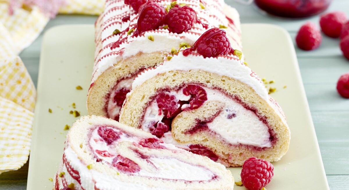 Eine sahnige Himbeerrolle mit Schmand liegt auf einer rechteckigen Porzellanplatte. Drumherum liegen Himbeeren und Servietten.