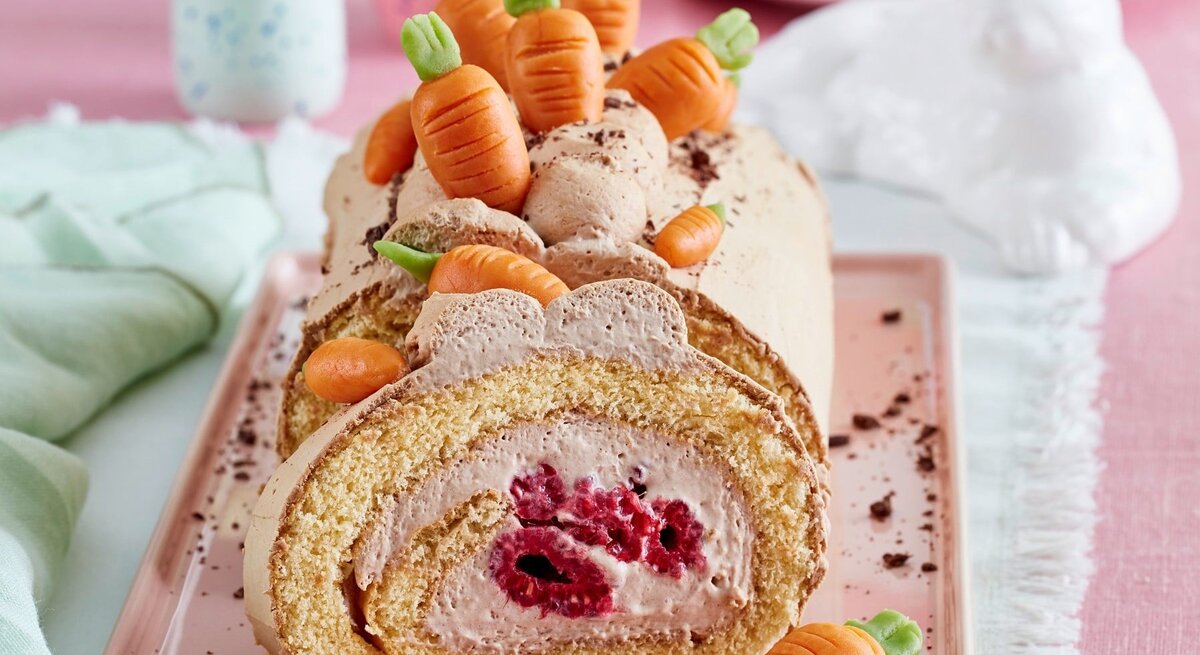 Auf einer Kuchenplatte ist eine Möhren-Biskuitrolle serviert. Sie ist mit Möhren aus Marzipan dekoriert. 