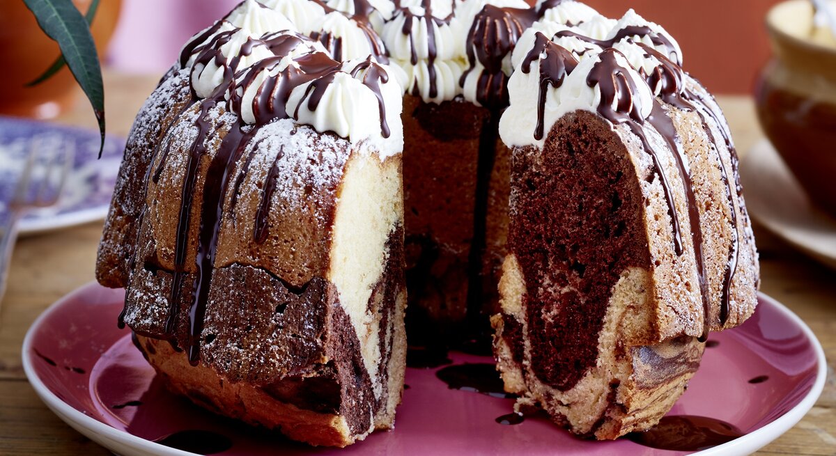 Auf einem Teller liegt ein angeschnittener Baileys-Marmorkuchen. Er ist mit Sahnetüpfeln verziert und mit Schokolade überzogen. 