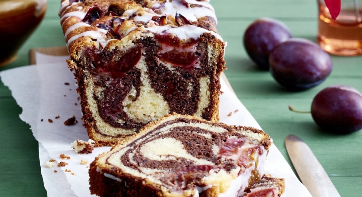 Auf einem Schneidebrett steht ein Zwetschgen Marmorkuchen. Er ist mit Puderzuckerguss überzogen. 