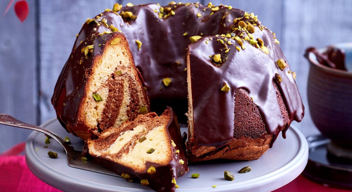 Ein angeschnittener Marmorkuchen auf einer Tortenplatte. Auf dem Tisch ist eine rote Tischdecke und dahinter eine rustikale Holzwand.