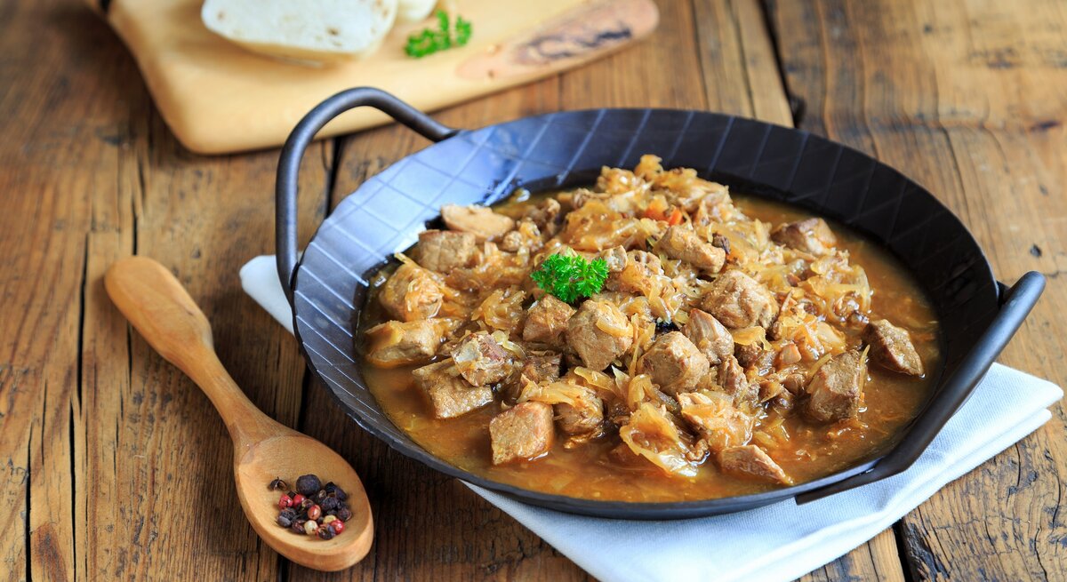 Frontalsicht: Eine schwarze, gusseiserne Pfanne mit Szegediner Gulasch liegt auf einem weißen Tuch, dass auf einem Holztisch liegt. Daneben liegen ein Holzlöffel mit Pfefferkörnernn. Und im Hintergrund sind aufgeschnittene Knödel.