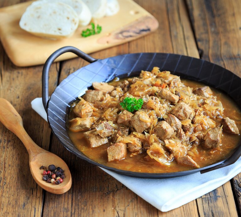 Frontalsicht: Eine schwarze, gusseiserne Pfanne mit Szegediner Gulasch liegt auf einem weißen Tuch, dass auf einem Holztisch liegt. Daneben liegen ein Holzlöffel mit Pfefferkörnernn. Und im Hintergrund sind aufgeschnittene Knödel. | © Shutterstock / juefraphoto