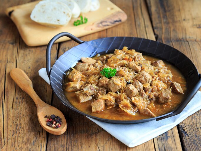 Frontalsicht: Eine schwarze, gusseiserne Pfanne mit Szegediner Gulasch liegt auf einem weißen Tuch, dass auf einem Holztisch liegt. Daneben liegen ein Holzlöffel mit Pfefferkörnernn. Und im Hintergrund sind aufgeschnittene Knödel. | © Shutterstock / juefraphoto