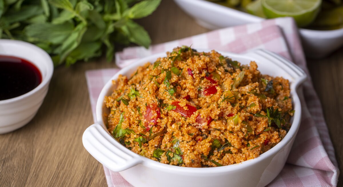 Frontalsicht: Auf einem Holztisch steht eine Schüssel Kisir, einen türkischen Bulgur-Salat. Dahinter liegen frische Minze, Zitronen und eine Schale mit einer roten Flüssigeit.