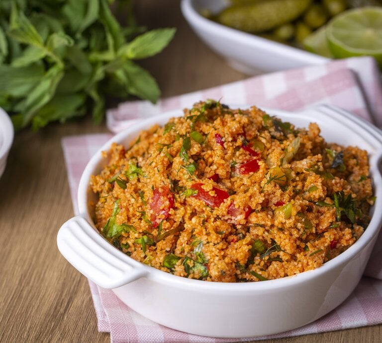 Frontalsicht: Auf einem Holztisch steht eine Schüssel Kisir, einen türkischen Bulgur-Salat. Dahinter liegen frische Minze, Zitronen und eine Schale mit einer roten Flüssigeit. | © Getty Images / Esin Deniz