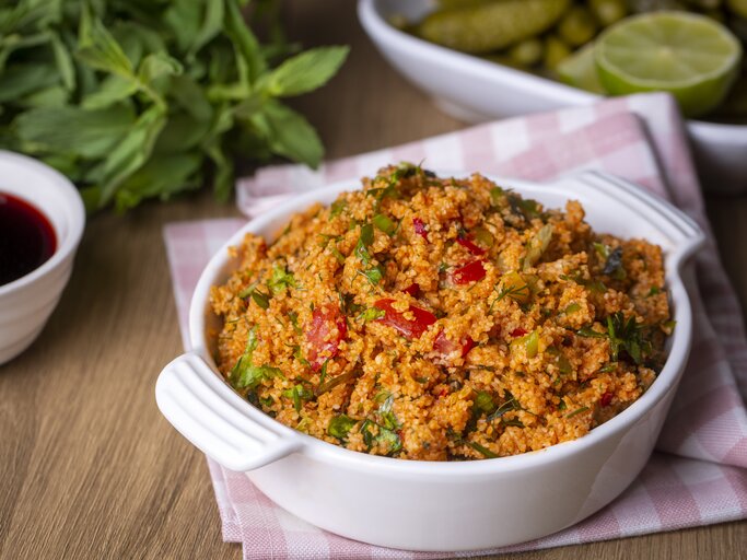 Frontalsicht: Auf einem Holztisch steht eine Schüssel Kisir, einen türkischen Bulgur-Salat. Dahinter liegen frische Minze, Zitronen und eine Schale mit einer roten Flüssigeit. | © Getty Images / Esin Deniz