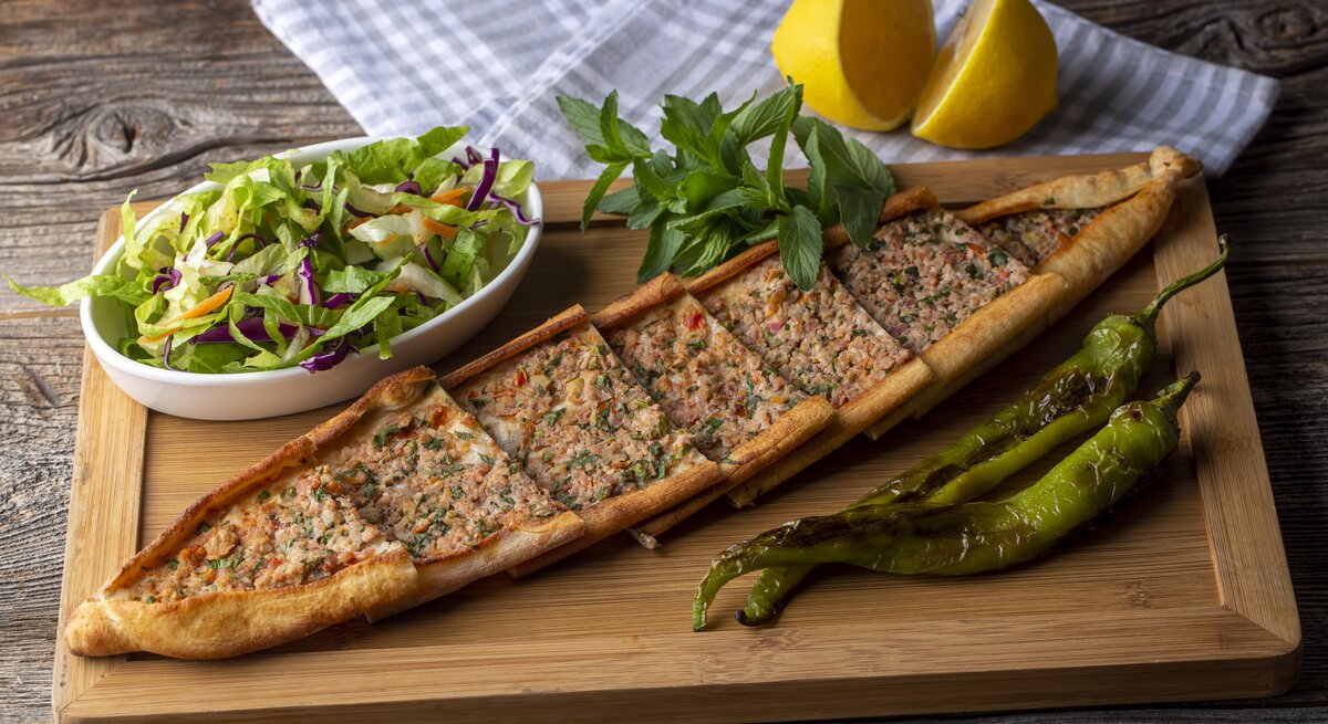 Ein Holzbrett, auf dem eine Pide mit Hackfleisch in Stücke geschnitten wurde. Daneben steht eine Schale Salat, außerdem liegen daneben eine aufgeschnittene Zitrone, ein Geschirrtuch, grüne Chilis und Kräuter.