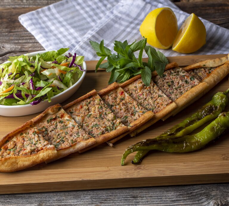 Ein Holzbrett, auf dem eine Pide mit Hackfleisch in Stücke geschnitten wurde. Daneben steht eine Schale Salat, außerdem liegen daneben eine aufgeschnittene Zitrone, ein Geschirrtuch, grüne Chilis und Kräuter. | © Getty Images/ Esin Deniz