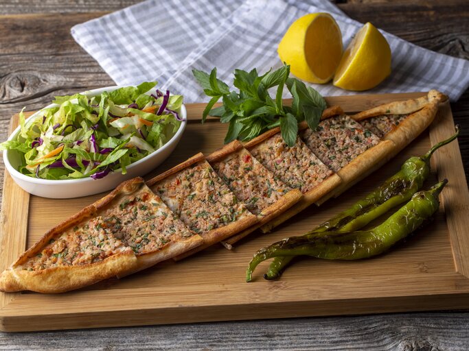 Ein Holzbrett, auf dem eine Pide mit Hackfleisch in Stücke geschnitten wurde. Daneben steht eine Schale Salat, außerdem liegen daneben eine aufgeschnittene Zitrone, ein Geschirrtuch, grüne Chilis und Kräuter. | © Getty Images/ Esin Deniz