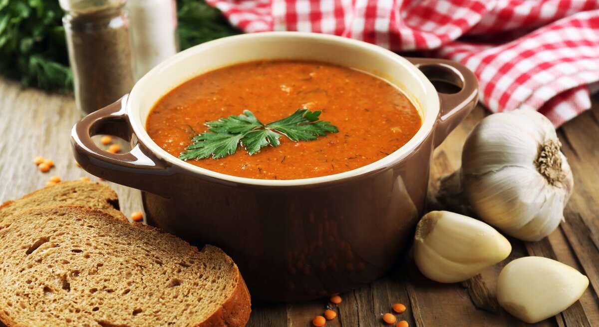 Frontalansicht: Auf einem Tisch steht eine Schale mit einer roten Suppe, die mit einem Blatt Petersilie garniert ist. Drumherum liegen Knoblauchzehen, eine Scheibe Brot und trockene rote Linsen.