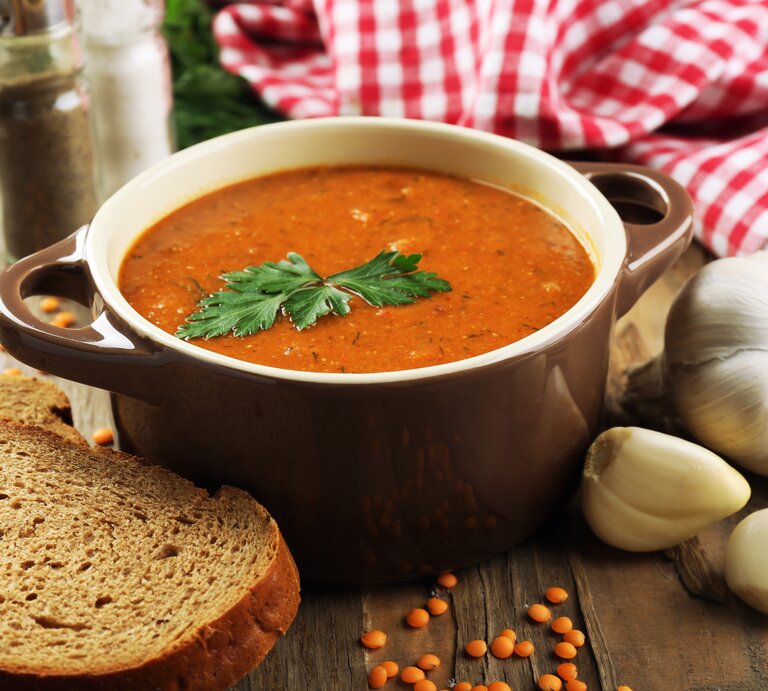Frontalansicht: Auf einem Tisch steht eine Schale mit einer roten Suppe, die mit einem Blatt Petersilie garniert ist. Drumherum liegen Knoblauchzehen, eine Scheibe Brot und trockene rote Linsen. | © Shutterstock / Africa Studio