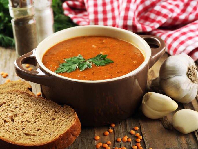 Frontalansicht: Auf einem Tisch steht eine Schale mit einer roten Suppe, die mit einem Blatt Petersilie garniert ist. Drumherum liegen Knoblauchzehen, eine Scheibe Brot und trockene rote Linsen. | © Shutterstock / Africa Studio