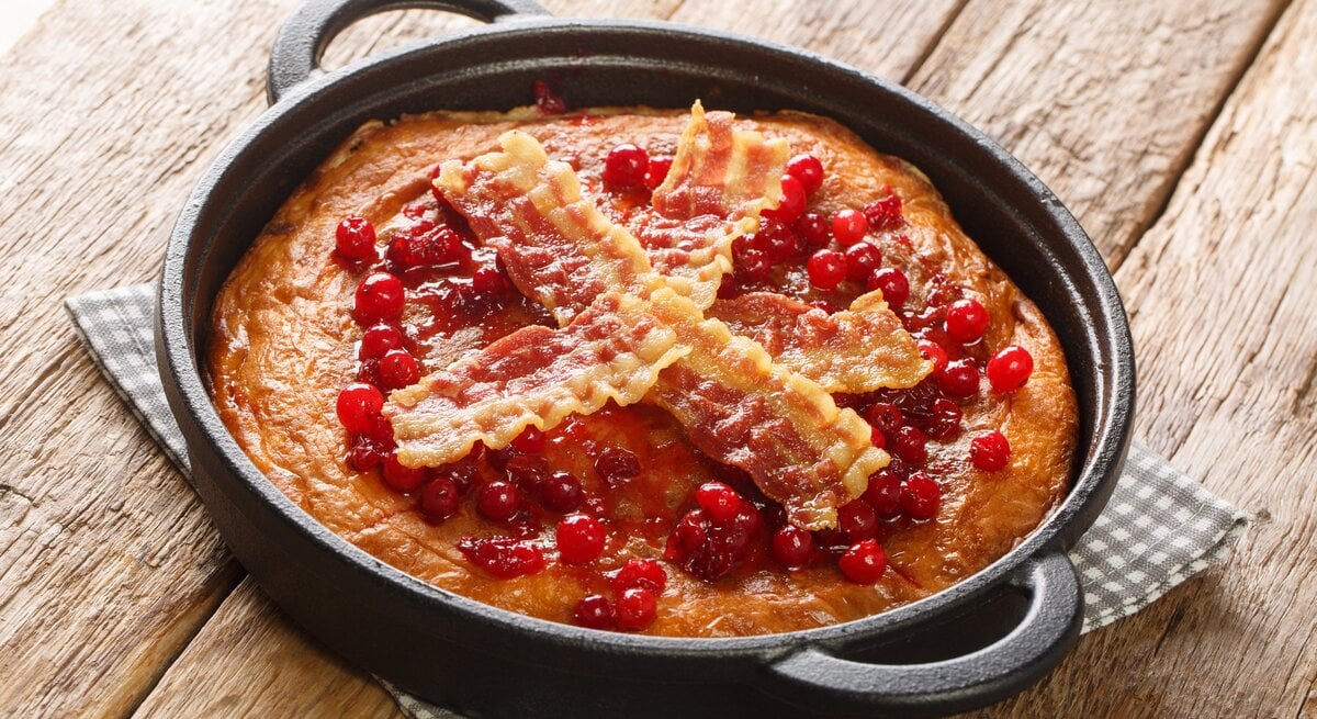 Eine Pfanne mit dem Äggakaka, dem schwedischen Eierkuchen garniert mit kross gebratenen Bacon-Scheiben und Preiselbeeren, auf einem karierten Untersetzer vor einem Holzhintergrund. 