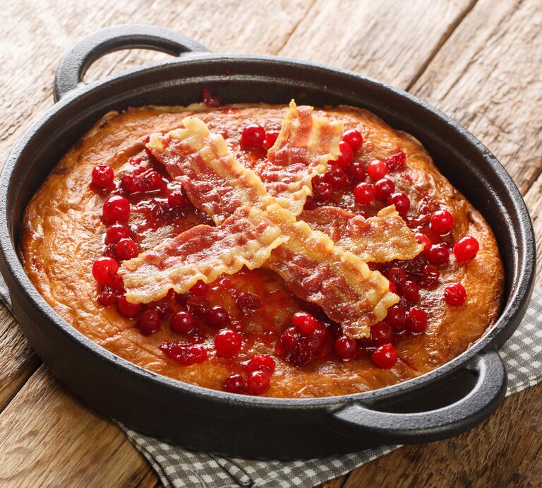 Eine Pfanne mit dem Äggakaka, dem schwedischen Eierkuchen garniert mit kross gebratenen Bacon-Scheiben und Preiselbeeren, auf einem karierten Untersetzer vor einem Holzhintergrund.  | © Getty Images/ ALLEKO