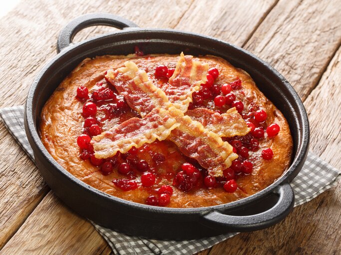 Eine Pfanne mit dem Äggakaka, dem schwedischen Eierkuchen garniert mit kross gebratenen Bacon-Scheiben und Preiselbeeren, auf einem karierten Untersetzer vor einem Holzhintergrund.  | © Getty Images/ ALLEKO