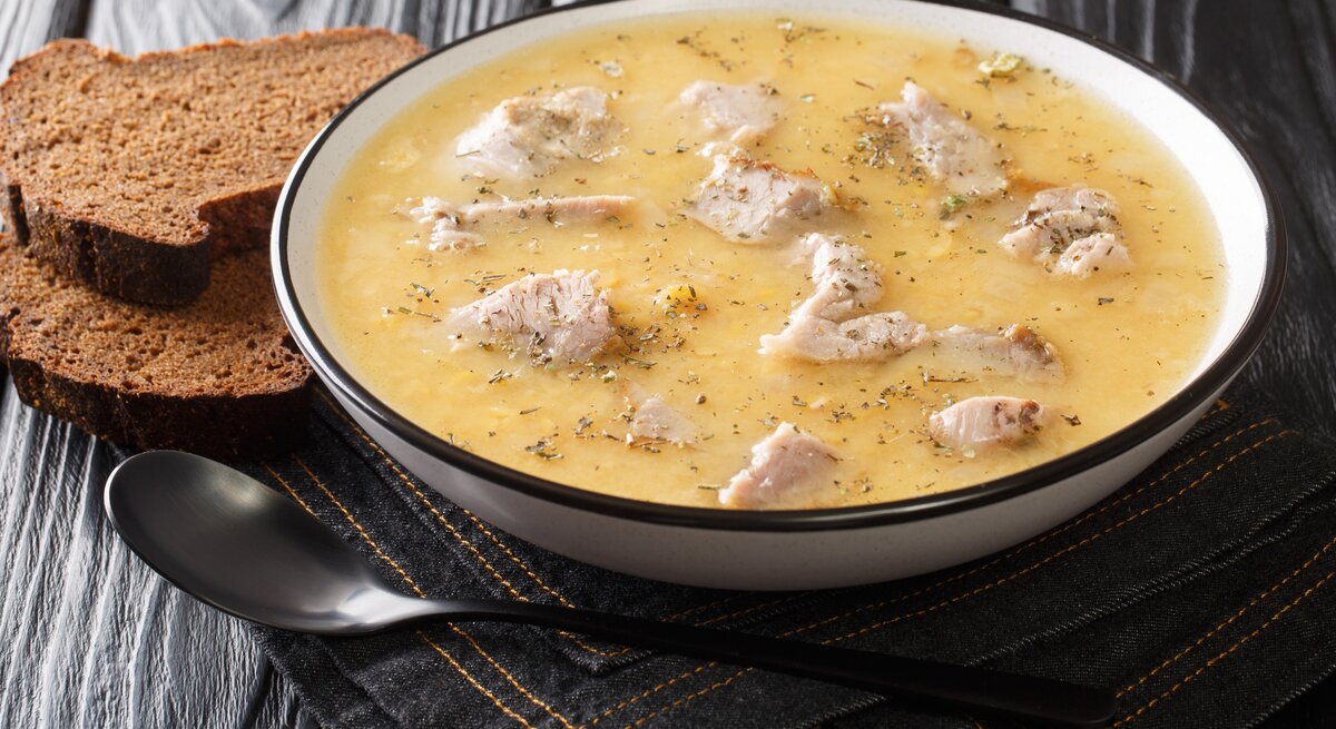 Ein voller Suppenteller mit der schwedischen Erbsensuppe mit Schweinefleisch, die Ärtsoppa neben einem Löffel und zwei Scheiben Schwarzbrot vor einem dunklen Hintergrund. 
