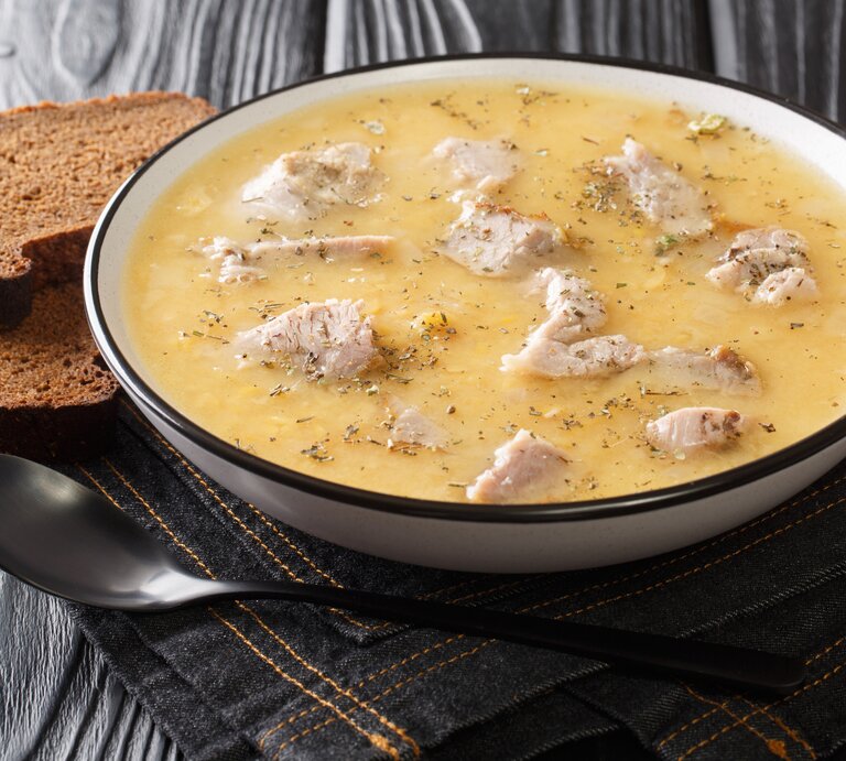 Ein voller Suppenteller mit der schwedischen Erbsensuppe mit Schweinefleisch, die Ärtsoppa neben einem Löffel und zwei Scheiben Schwarzbrot vor einem dunklen Hintergrund.  | © Getty Images/ FomA