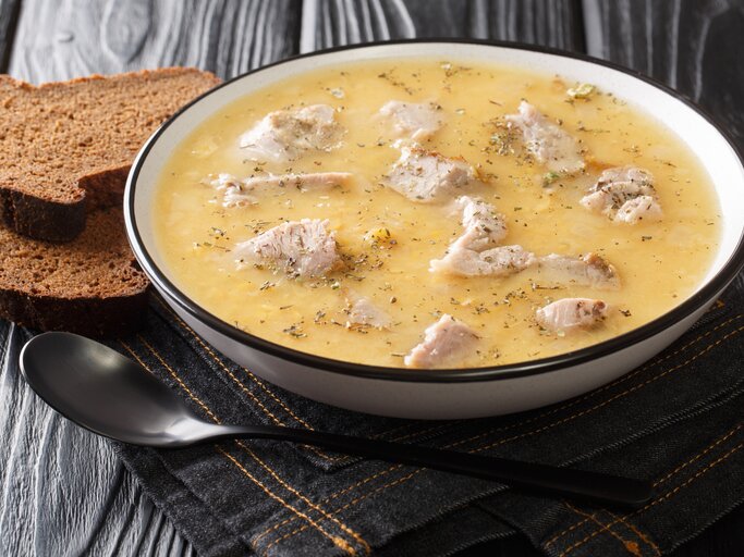 Ein voller Suppenteller mit der schwedischen Erbsensuppe mit Schweinefleisch, die Ärtsoppa neben einem Löffel und zwei Scheiben Schwarzbrot vor einem dunklen Hintergrund.  | © Getty Images/ FomA