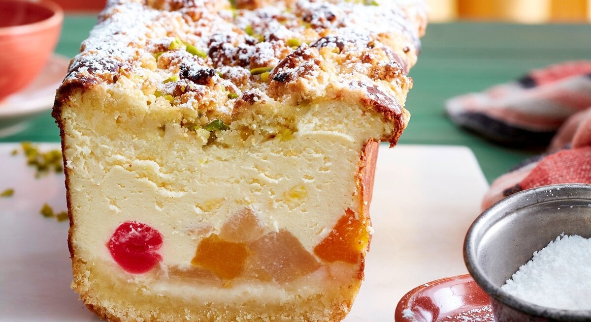 Auf einem Brett steht ein Tutti-Frutti-Käsekuchen aus der Kastenform. Er ist mit Puderzucker bestreut.