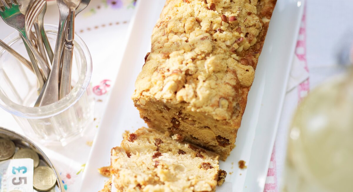 Apfel-Kastenkuchen mit Streuseln angeschnitten und serviert auf einem länglichen weißem Teller und hellem Untergrund.