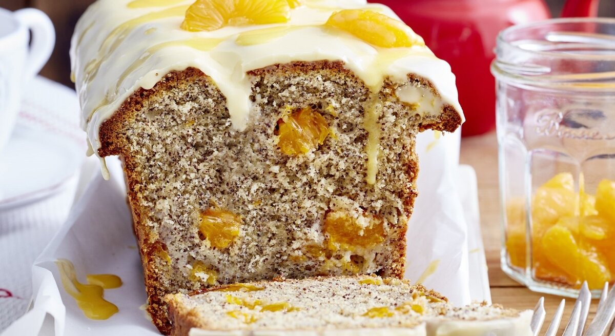 Auf einem Tisch steht ein angeschnittener Mohn-Eierlikör-Kastenkuchen. Er ist mit Mandarinen bedeckt. 