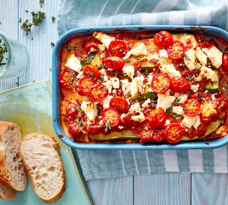 Eine Zucchini-Lasagne mit Feta in einer blauen Auflaufform steht auf einem Geschirrtuch auf einem blauen Tisch. Die Lasagne ist angerichtet mit Feta, Tomaten und Thymian. Neben der Auflaufform liegt Baguette und ein Glas mit Kräutern. | © EatClub, Isabell Triemer