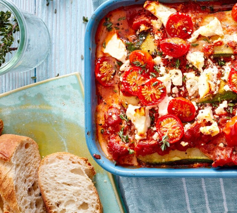 Eine Zucchini-Lasagne mit Feta in einer blauen Auflaufform steht auf einem Geschirrtuch auf einem blauen Tisch. Die Lasagne ist angerichtet mit Feta, Tomaten und Thymian. Neben der Auflaufform liegt Baguette und ein Glas mit Kräutern. | © EatClub, Isabell Triemer
