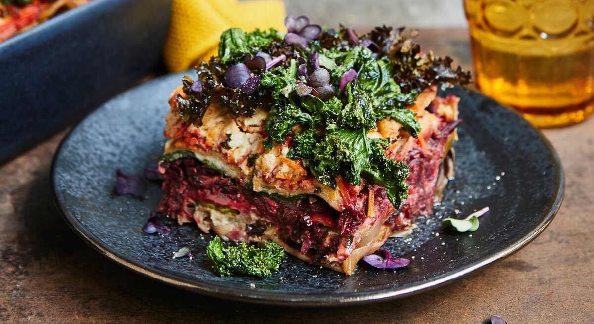 Eine Portion bunte Winterlasagne mit Grünkohl und rote Bete steht auf einem dunklen Teller auf einem Holztisch. Die Lasagne ist mit bunter Kresse angerichtet. Im Hintergrund steht die Ofenform mit der restlichen Lasagne und ein Glas.