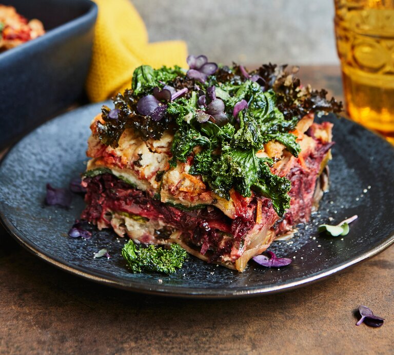 Eine Portion bunte Winterlasagne mit Grünkohl und rote Bete steht auf einem dunklen Teller auf einem Holztisch. Die Lasagne ist mit bunter Kresse angerichtet. Im Hintergrund steht die Ofenform mit der restlichen Lasagne und ein Glas. | © StockFood Studios / Sporrer, Brigitte