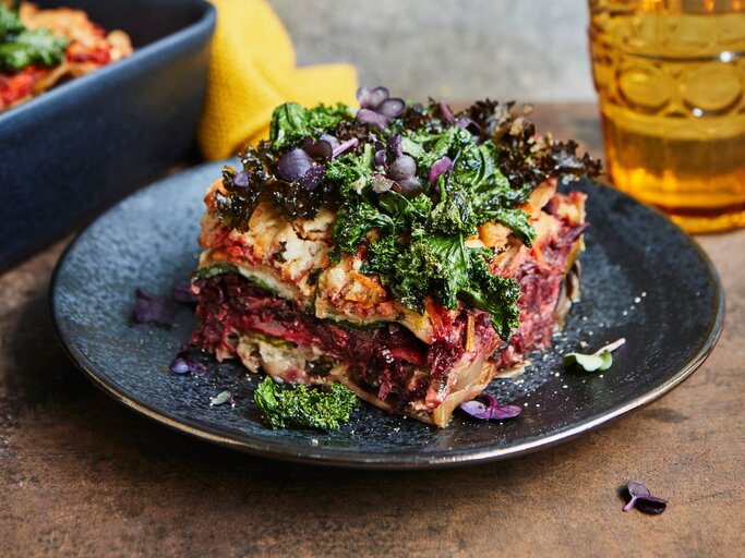 Eine Portion bunte Winterlasagne mit Grünkohl und rote Bete steht auf einem dunklen Teller auf einem Holztisch. Die Lasagne ist mit bunter Kresse angerichtet. Im Hintergrund steht die Ofenform mit der restlichen Lasagne und ein Glas. | © StockFood Studios / Sporrer, Brigitte