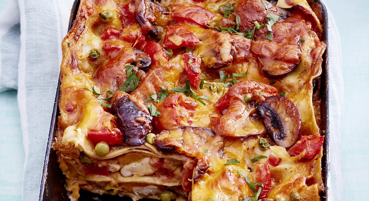 Hähnchen-Lasagne mit Pilzen und frischen Tomaten, angeschnitten in einer großen Auflaufform auf einem weißen Tuch serviert. 