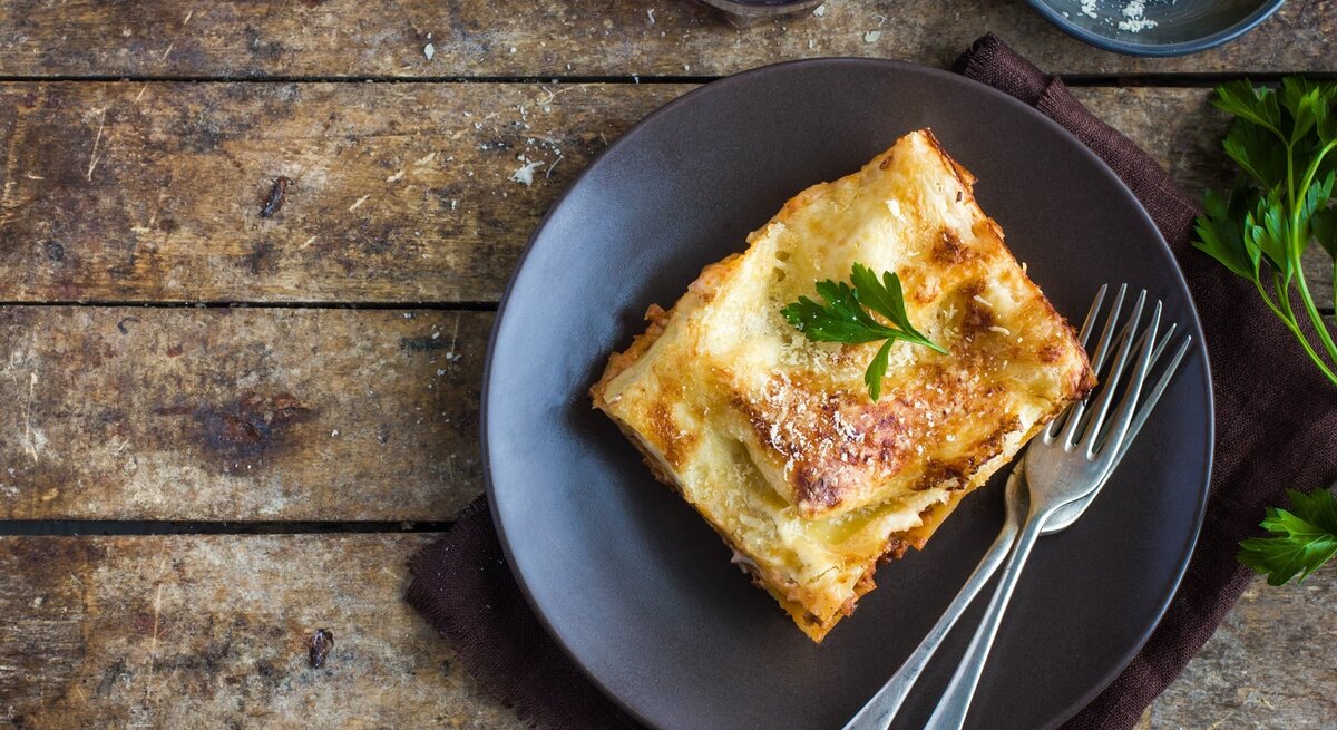 Ein Stück Lasagne auf einem rustikalen Holztisch von oben fotografiert. Darüber eine kleine Schale mit geriebenem Käse und ein Glas mit Rotwein.
