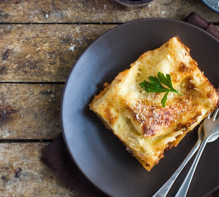 Ein Stück Lasagne auf einem rustikalen Holztisch von oben fotografiert. Darüber eine kleine Schale mit geriebenem Käse und ein Glas mit Rotwein. | © Shutterstock/ Anna Shepulova