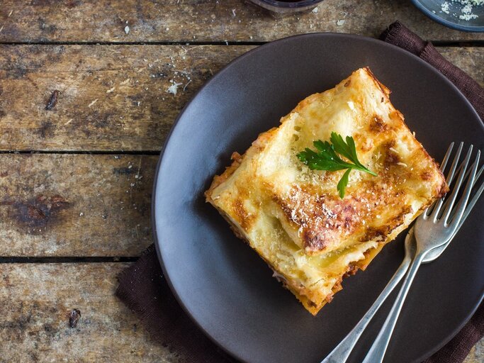 Ein Stück Lasagne auf einem rustikalen Holztisch von oben fotografiert. Darüber eine kleine Schale mit geriebenem Käse und ein Glas mit Rotwein. | © Shutterstock/ Anna Shepulova