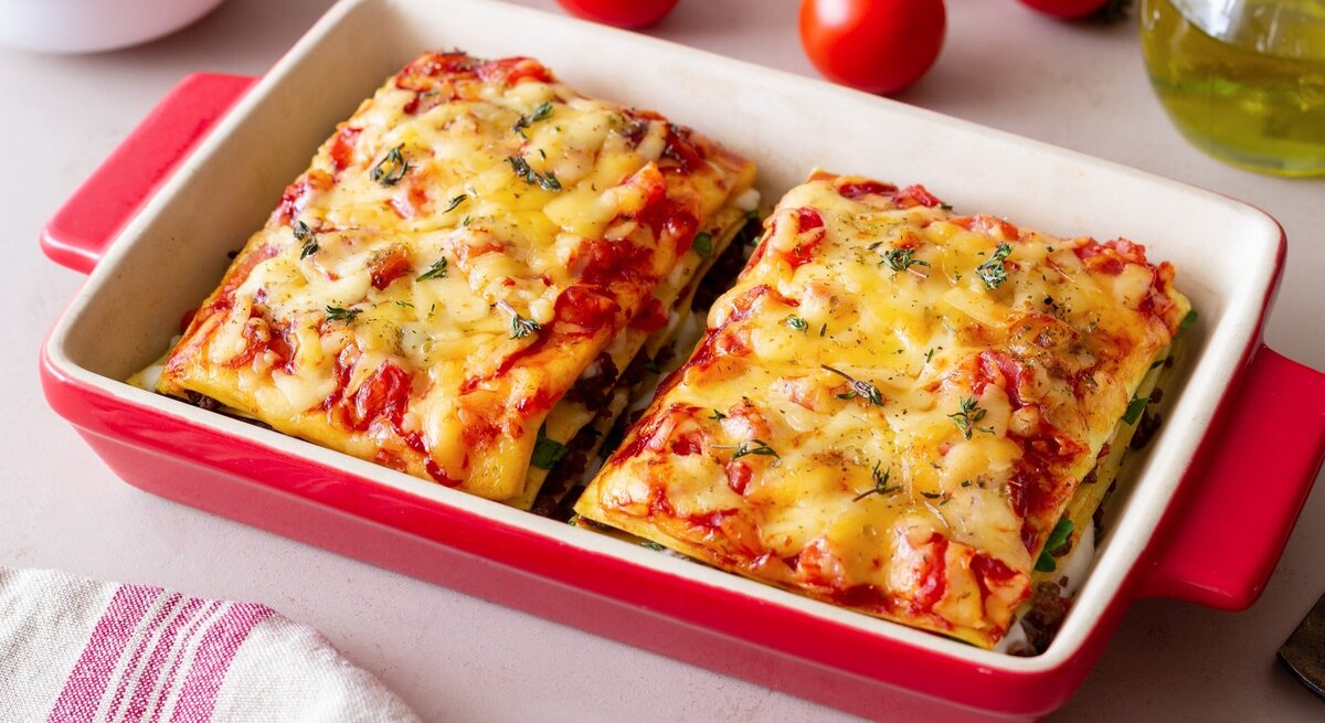 Bärlauch-Lasagne in roter Auflaufform mit Tomaten auf hellen Tisch. Rechts Flasche mit Olivenöl, links Schale mit Parmesan und Cocktailtomaten.