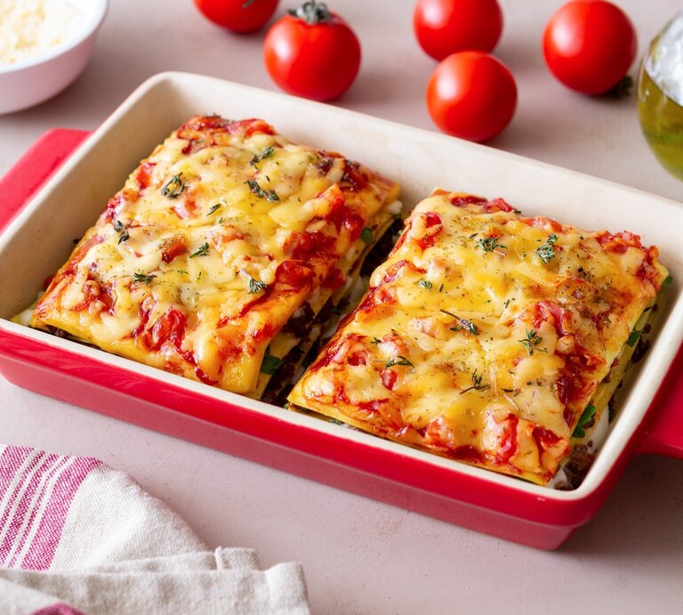Bärlauch-Lasagne in roter Auflaufform mit Tomaten auf hellen Tisch. Rechts Flasche mit Olivenöl, links Schale mit Parmesan und Cocktailtomaten. | © Getty Images / Vladislav Chusov