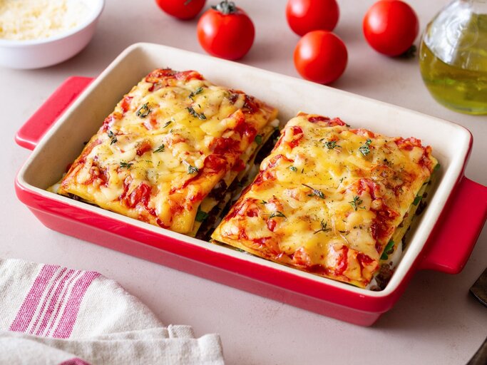 Bärlauch-Lasagne in roter Auflaufform mit Tomaten auf hellen Tisch. Rechts Flasche mit Olivenöl, links Schale mit Parmesan und Cocktailtomaten. | © Getty Images / Vladislav Chusov