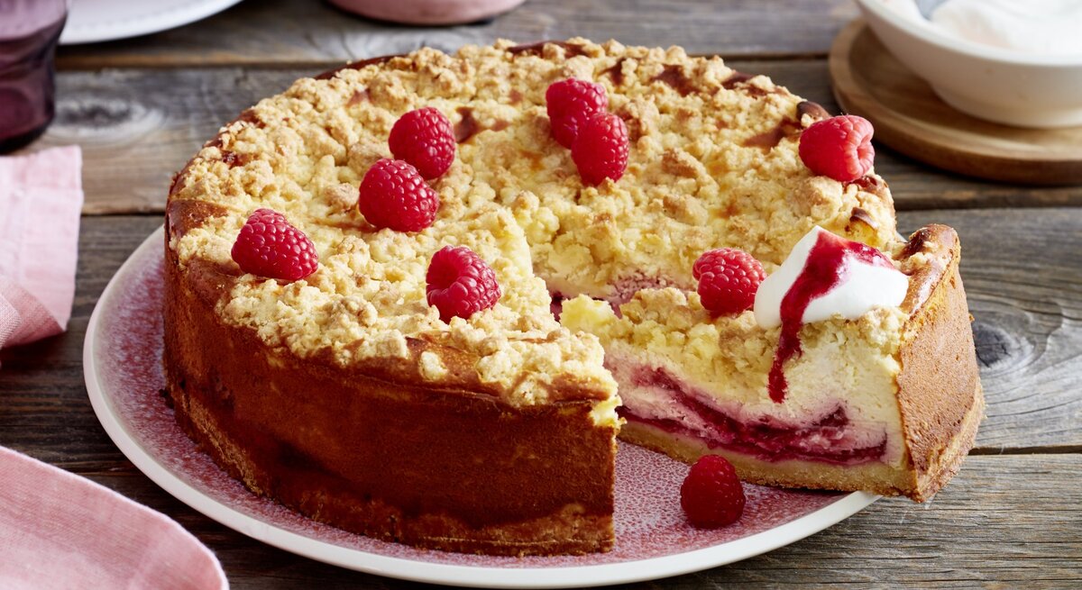 Angeschnittener Himbeer-Käsekuchen mit Sahne auf einem rosa Teller, im Hintergrund Geschirr und eine Schüssel mit Sahne, alles auf Holzuntergrund. 