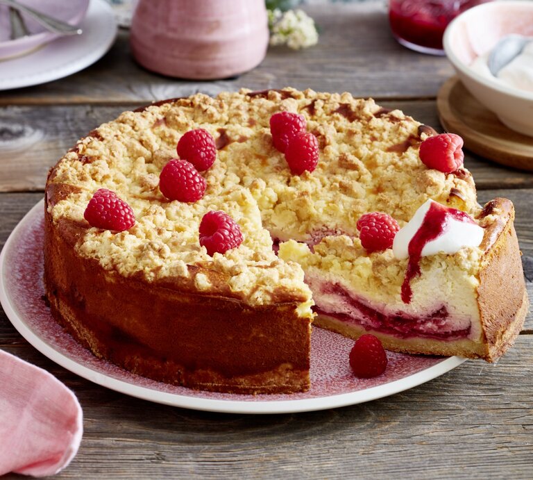 Angeschnittener Himbeer-Käsekuchen mit Sahne auf einem rosa Teller, im Hintergrund Geschirr und eine Schüssel mit Sahne, alles auf Holzuntergrund.  | © EAT CLUB / Isabell Triemer 