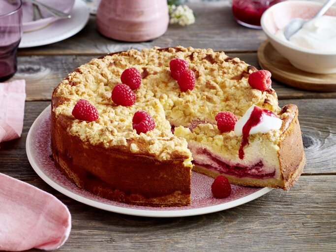 Angeschnittener Himbeer-Käsekuchen mit Sahne auf einem rosa Teller, im Hintergrund Geschirr und eine Schüssel mit Sahne, alles auf Holzuntergrund.  | © EAT CLUB / Isabell Triemer 
