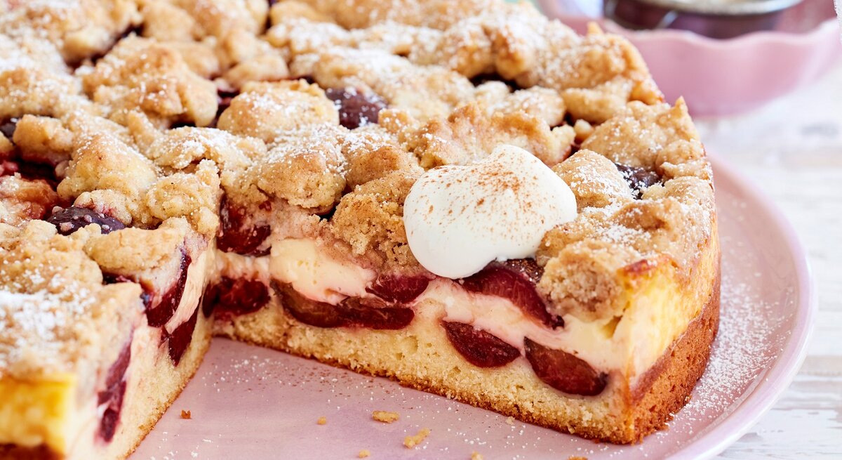 Pflaumen-Puddingkuchen mit Zimtstreuseln, angeschnitten und bestreut mit Puderzucker und serviert mit geschlagener, süßer Sahne auf einem rosa Tisch. 