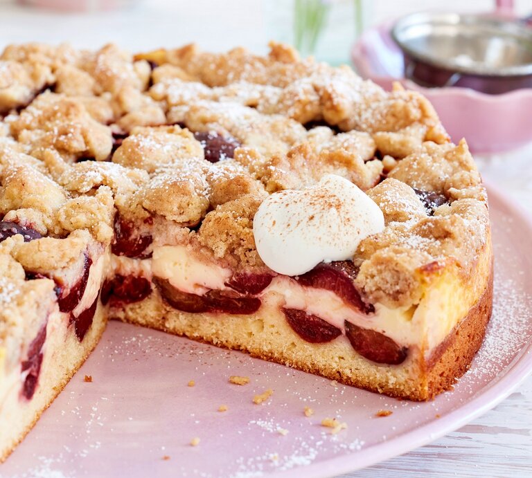 Pflaumen-Puddingkuchen mit Zimtstreuseln, angeschnitten und bestreut mit Puderzucker und serviert mit geschlagener, süßer Sahne auf einem rosa Tisch.  | © EAT CLUB / Isabell Triemer